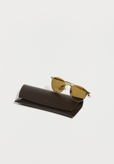 Στρογγυλά γυαλιά ηλίου με χρυσή κορνίζα και φακούς με καφέ απόχρωση, τοποθετημένα πάνω σε κλειστή θήκη γυαλιών από σκούρο καφέ δέρμα.