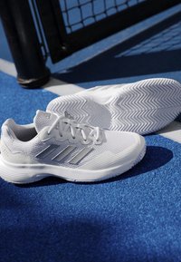 Pair of white Adidas athletic shoes with silver stripes placed on blue sports court near black net post.