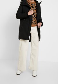 Chaqueta negra con cremallera y capucha sobre un suéter marrón con estampado de leopardo, combinada con pantalones anchos de pana blancos y botines blancos.