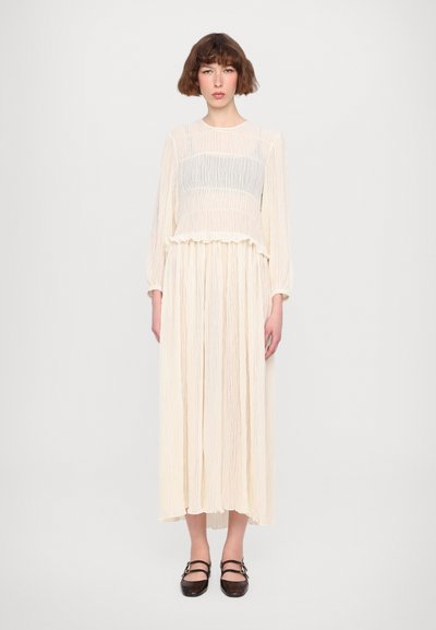 Robe longue crème claire avec une texture plissée, présentant un haut plus court et froncé ainsi que des manches longues. Le mannequin porte des chaussures plates marron foncé.