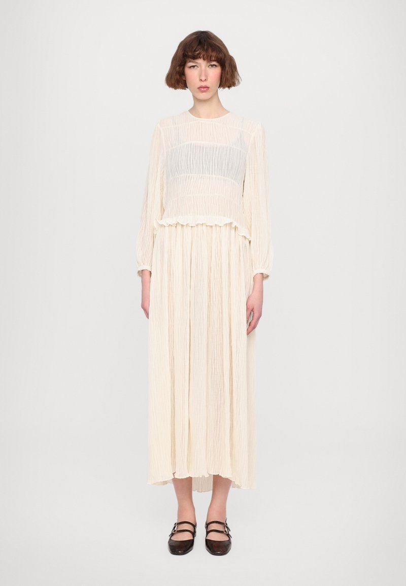 Long, light cream dress with a pleated texture, featuring a shorter, gathered top and long sleeves. The model wears dark brown, flat shoes.