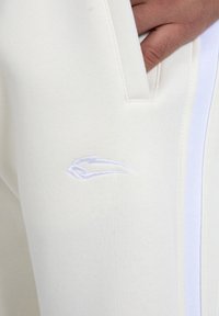Valkoiset puuvillajoggersit, joissa on sivutasku, valkoinen raita sivussa ja vaaleansinisesti broderattu logo reidessä.