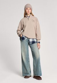 Beżowy polarowy pulower z półzamkiem, niebieska w kratkę flanela poniżej, jasnoniebieskie szerokie dżinsy i brązowe klapki, na głowie z wełnianą czapką.