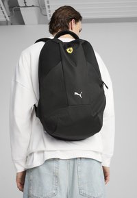 Sac à dos Puma noir avec panneaux en maille, logo Ferrari et branding Puma blanc. Le design comprend des bretelles ajustables et une fermeture éclair.