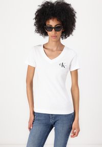 White cotton V-neck t-shirt featuring a small black logo on the left chest, paired with blue denim jeans and black angular sunglasses.