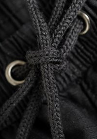 Schwarzer Stoff mit einem strukturierten Muster, verflochtene schwarze Kordelspitzen, die durch silberne Metallösen gefädelt sind und zu einem Knoten gebunden werden.