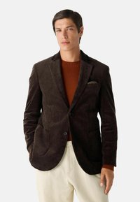 Bruine corduroy blazer met notch lapels, twee zakken en een knoopsluiting, gelaagd over een roestkleurige trui, gecombineerd met lichte broeken.