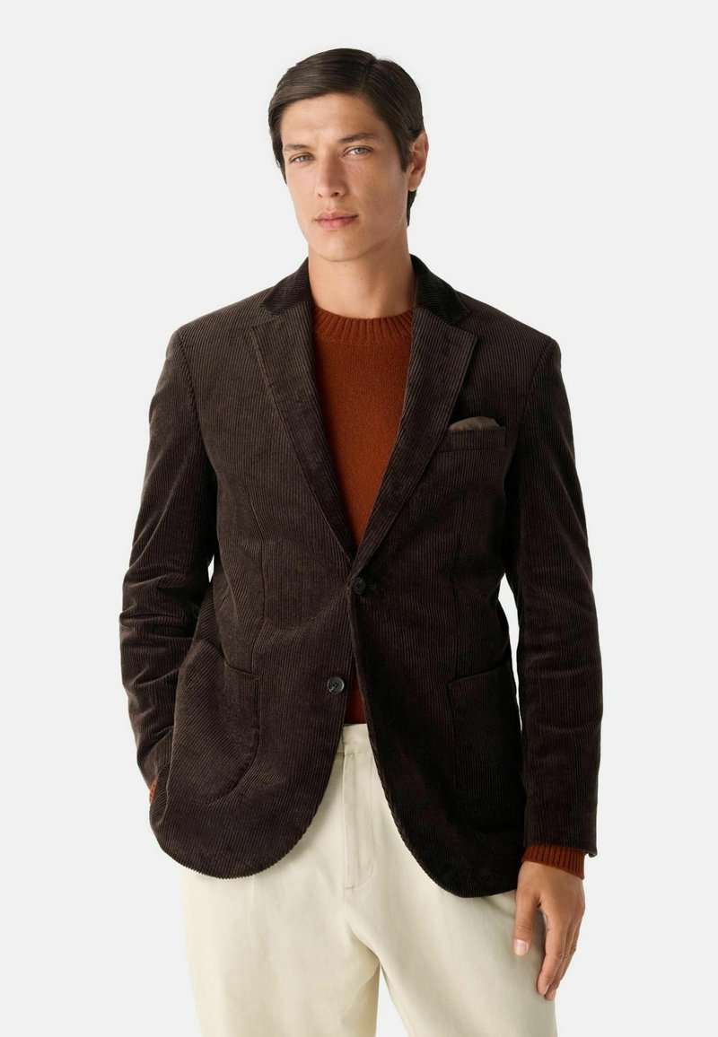Bruine corduroy blazer met notch lapels, twee zakken en een knoopsluiting, gelaagd over een roestkleurige trui, gecombineerd met lichte broeken.