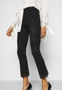 Zwarte fitted broek met kantafwerking aan de onderkant, voorzien van geborduurde bloemenpatronen in een donkerder tint, gecombineerd met een lichte blouse.