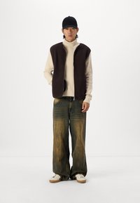 Brauner Fleece-Zip-Weste über einem cremefarbenen Rollkragenpullover, kombiniert mit weit geschnittenen Jeanshosen mit strukturiertem Finish. Weiße Sneaker runden den Look ab.