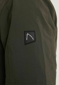 Ciemnozielona tkanina o gładkiej fakturze z czarną naszywką z białym geometrycznym wzorem, umieszczoną na górnej części rękawa.