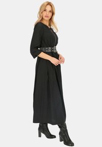 Czarna długa sukienka z długim rękawem i podkreśloną talią, z szerokim paskiem ze srebrnymi oczkami. Modelka nosi czarne botki na wysokim obcasie do kolan.