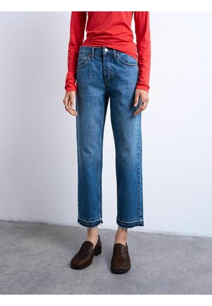 Kvinde iført cropped blå jeans med frynset kant, brune loafers og en rød bluse med lange ærmer, stående på en grå gulv mod en hvid væg.