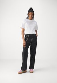 Witte katoenen t-shirt met een klein logo, gecombineerd met zwarte denim jeans met wijde pijpen en kettingdetails, gedragen met roze sneakers.