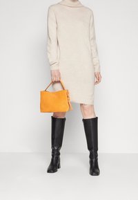 Abito a collo alto in maglia beige, lungo fino alla caviglia. Borsa in suede arancione con manico e dettaglio a fiocco. Stivali neri in pelle al ginocchio con tacco.