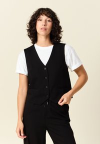 Svart väst med V-hals, fem knappar och två framfickor, bärs över en vit T-shirt, med en skräddarsydd passform och mjuk textur.