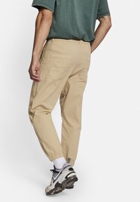 Beige cargobukser med elastiske ankellommer og flere baglommer, lavet af blødt bomuldsstof. Båret med en grøn skjorte og sneakers.