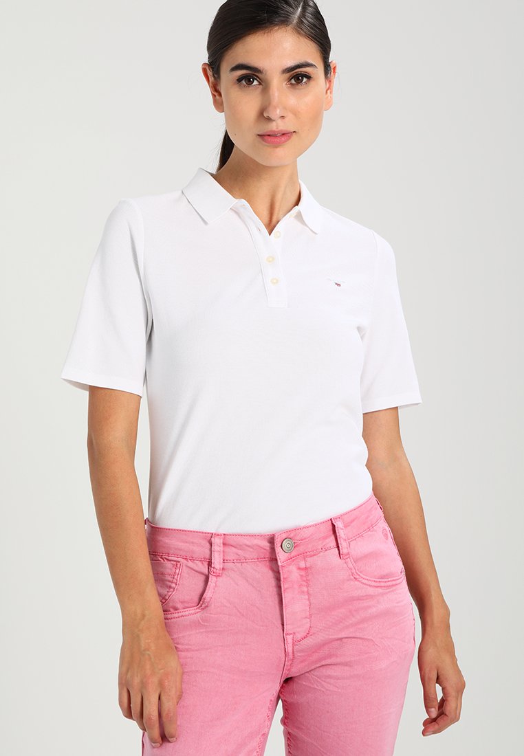 Weißes Polohemd mit Kragen und kurzen Ärmeln, mit einem kleinen gestickten Logo. Kombiniert mit rosa Jeans. Glatte Textur.