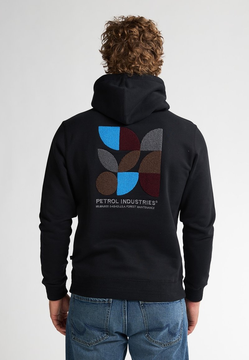 Personne aux cheveux bouclés portant un sweat à capuche noir avec des formes géométriques colorées et le texte « Petrol Industries » dans le dos, associé à un jean bleu.