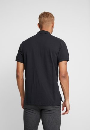 Mann mit kurzen, lockigen Haaren, der ein schwarzes, kurzärmliges Polohemd und eine Hose mit feinen Streifen trägt, von hinten vor einem einfarbigen Hintergrund stehend.