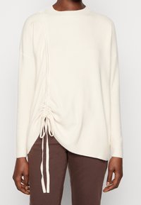 Pull tricoté couleur crème avec un col rond, un détail froncé sur le côté avec des liens, et des manches longues. Le tissu a une texture douce.