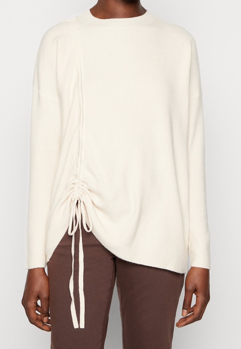 Pull tricoté couleur crème avec un col rond, un détail froncé sur le côté avec des liens, et des manches longues. Le tissu a une texture douce.