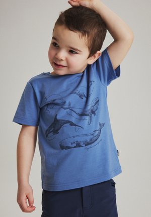 Jonge jongen die een blauw T-shirt draagt met gedetailleerde zwarte schetsen van walvissen en zeezoogdieren, terwijl hij één arm boven zijn hoofd heft.