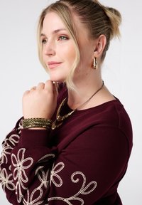 Bordeaux sweatshirt met witte geborduurde bloemenontwerpen op de mouw, gelaagde gouden kettingen en meerdere goudkleurige armbanden om de pols.