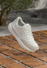 Valge Calvin Klein'i spordijalats, millel on paelkinnitus, tekstureeritud nahk ja sametised lisandused, eksponeeritud väljas puidust ruudukujuliste plaatide kohal.