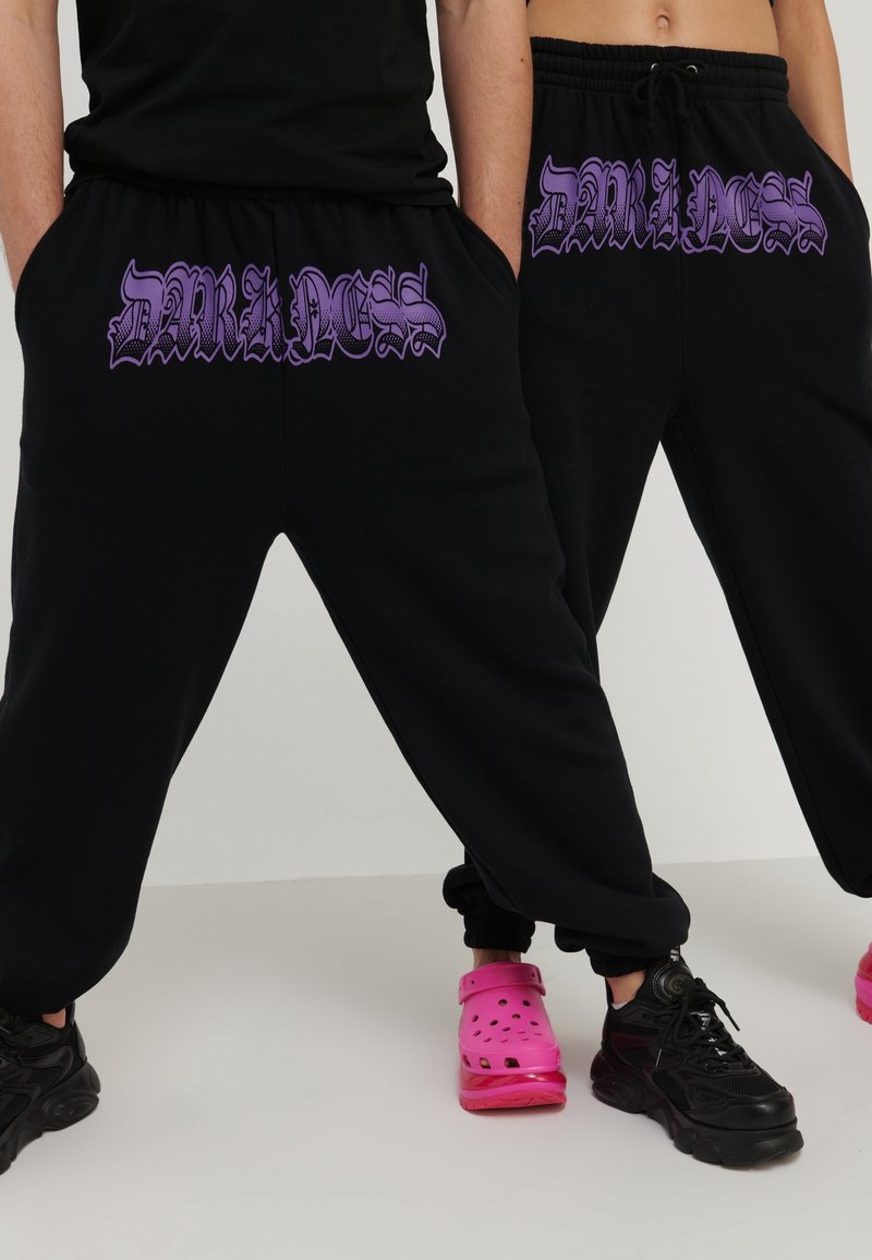 Svarta svettbyxor med lila textgrafik "DARKNESS" på framsidan. Har elastiska muddar och midjeband med dragsko.