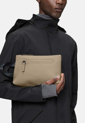 Man in een zwarte jas houdt een beige RAINS etui met een voorvak met rits vast tegen een effen achtergrond.