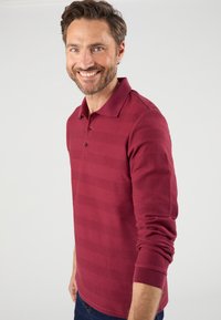 Bordeaux gestreepte polo met een ribbelstructuur, drie-knops sluiting en lange mouwen, casual gedragen door een glimlachende man.