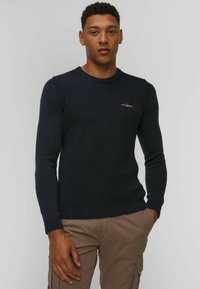 Maglione navy con texture a coste e scollo tondo. Presenta maniche lunghe e un piccolo logo dorato sul petto. Indossato con pantaloni beige.