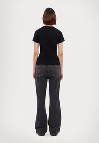 En person som bär en svart kortärmad skjorta och lösa svarta jeans, står med ryggen vänd framåt mot en enkel vit bakgrund.