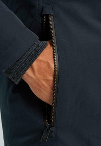 Giacca navy con una tasca con zip. I denti della cerniera dorata contrastano con il tessuto scuro. Il polsino presenta una texture a coste grigia.