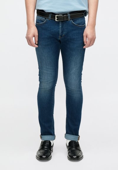 Jean skinny bleu légèrement délavé, en denim, design à cinq poches, assorti à des chaussures noires sans lacets et une ceinture noire.
