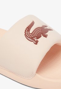 Sandale glissante crème avec une semelle texturée, ornée d'un logo crocodile brun sur la sangle. Matériau synthétique lisse avec un profil bas.