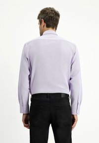 Długi rękaw, jasnofioletowa koszula z teksturowanej tkaniny i zapiętymi mankietami, w zestawieniu z czarnymi dżinsami. Widok z tyłu.