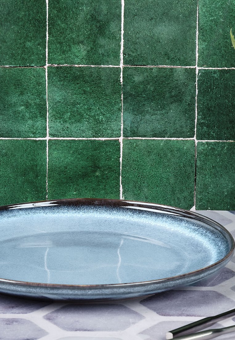 Piatto rotondo con una superficie lucida di un blu chiaro e un bordo scuro, posizionato su uno sfondo di piastrelle verdi. La texture è liscia.