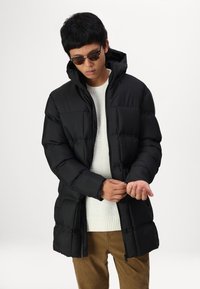 Schwarze Daunenjacke mit Kapuze, im kastigen Design, mit gesteppter Textur und Fronttaschen. Getragen über einem weißen gestrickten Pullover.