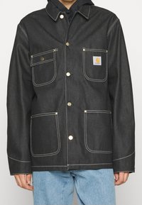 Chaqueta de mezclilla negra con dos bolsillos en el pecho, botones dorados, detalles de costura blanca y una etiqueta en el bolsillo izquierdo. Lleva puesta una sudadera con capucha negra.