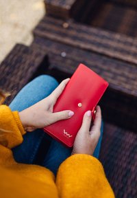 Red leather wallet with a smooth texture, featuring a gold snap button and embossed branding at the bottom corner. Hands holding the wallet.