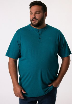 Türkises Henley-Shirt mit kurzen Ärmeln, drei schwarzen Knöpfen und einem subtilen Logo. Der Stoff ist glatt und der Schnitt ist lässig. Getragen mit dunklen Jeans.
