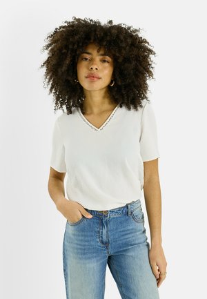 Jeune femme aux cheveux bouclés portant un haut blanc à col en V et manches courtes, ainsi qu'un jean bleu, debout avec une main dans la poche.