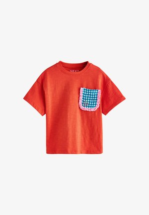 Piros rövid ujjú póló kék-fehér kockás mellzsebbel, amelyet rózsaszín fodrok díszítenek, valamint egy kis hímzett minta.