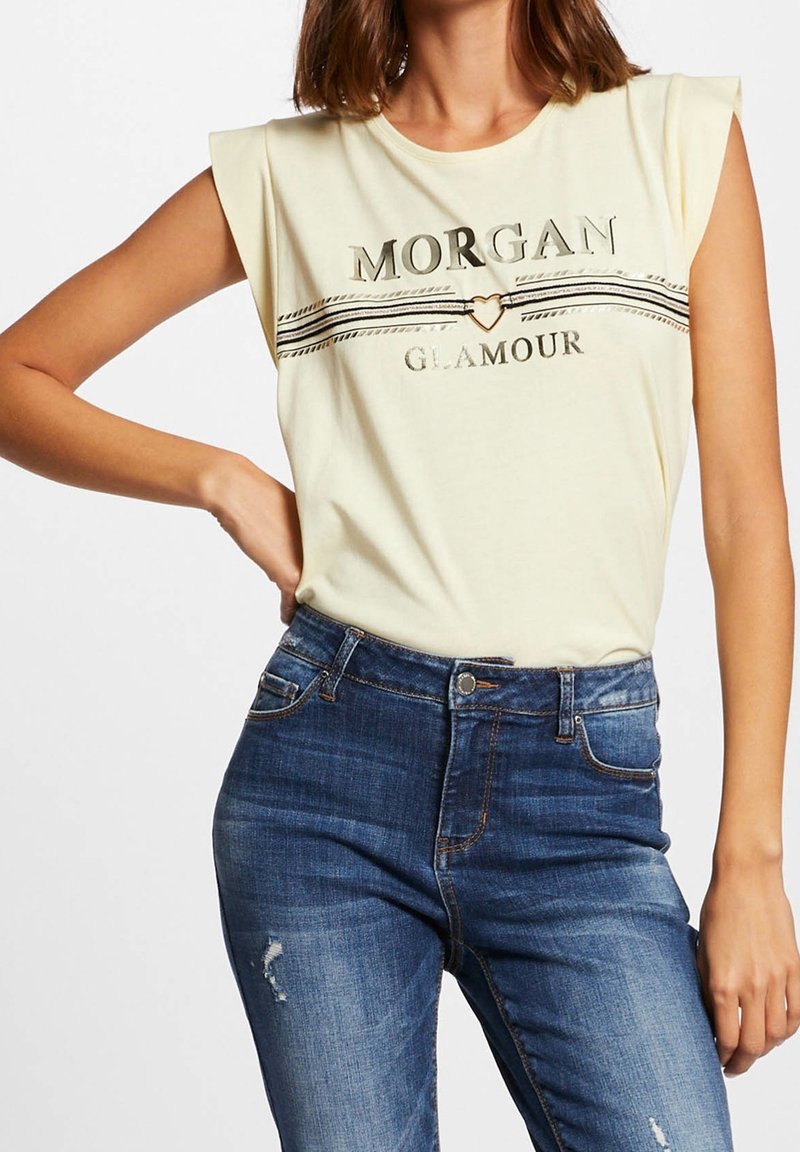 Femme portant un top sans manches crème avec le texte "Morgan Glamour" et un détail cœur, associé à un jean bleu taille haute légèrement usé.