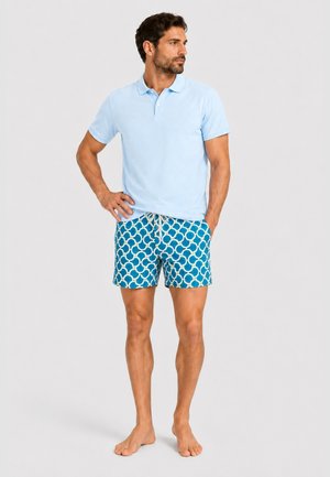 Mand stående barfodet iført en lyseblå polo shirt og blå mønstrede shorts med hvid snøre på en ensfarvet baggrund.