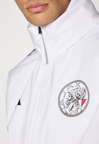Hvid jakke med h�øj krave, lynlåslukning og et broderet Ajax-logo med en klassisk figur. Glat tekstur, minimalistisk design.