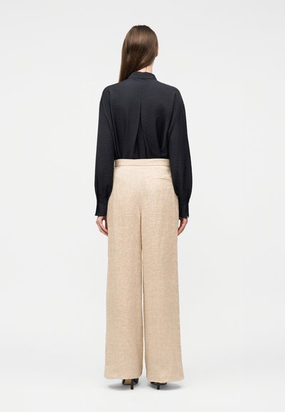 Γυναίκα με μακριά καστανά μαλλιά που στέκεται με την πλάτη στραμμένη, φοράει μαύρο πουκάμισο με μακριά μανίκια που είναι μπαλωμένο σε παντελόνι σε ευρεία γραμμή με ψηλή μέση σε μπεζ απόχρωση και μαύρες γόβες.