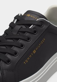 Κοντινή λήψη μαύρου αθλητικού παπουτσιού Tommy Hilfiger με μαύρα κορδόνια, χρυσό λογότυπο στο πλάι και λευκή σόλα.